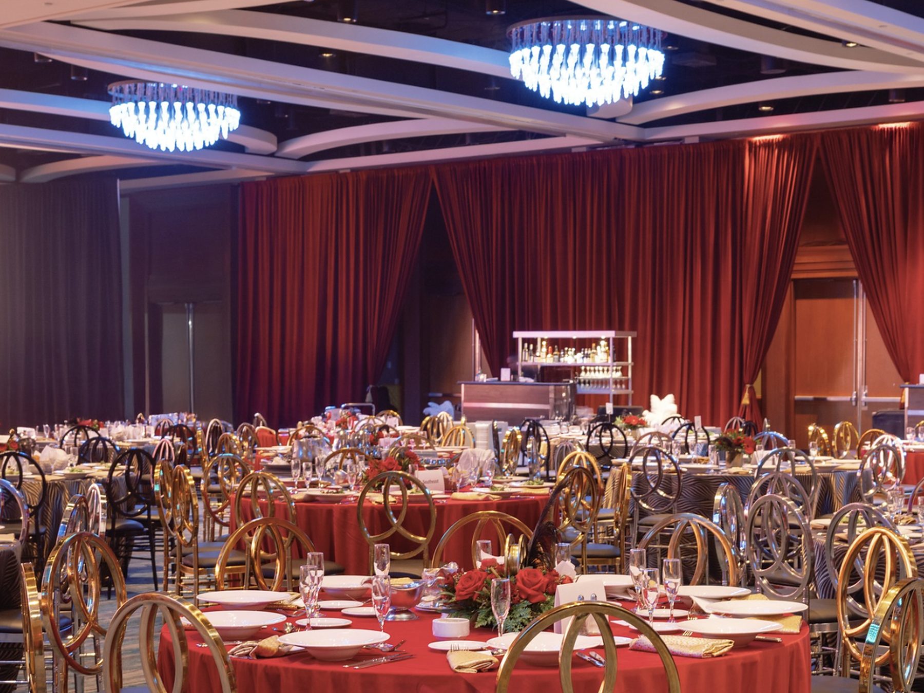 Ballroom banquet set with red round linens, gold ring-back chairs, red stage drapes, and crystal chandeliers